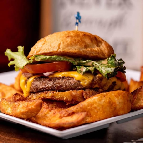 Cheeseburger with lettuce, tomato, and a blue toothpick, served with potato wedges on a white plate.
