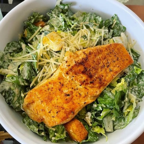 A white bowl filled with Caesar salad, topped with a large piece of grilled salmon and shredded cheese.